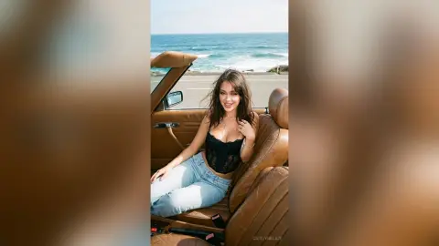 Smiling woman in a black corset and light blue jeans sitting in a vintage convertible by the ocean.