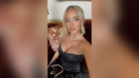 Glamorous blonde woman in a black satin dress and pearl necklace sitting on a vintage sofa.