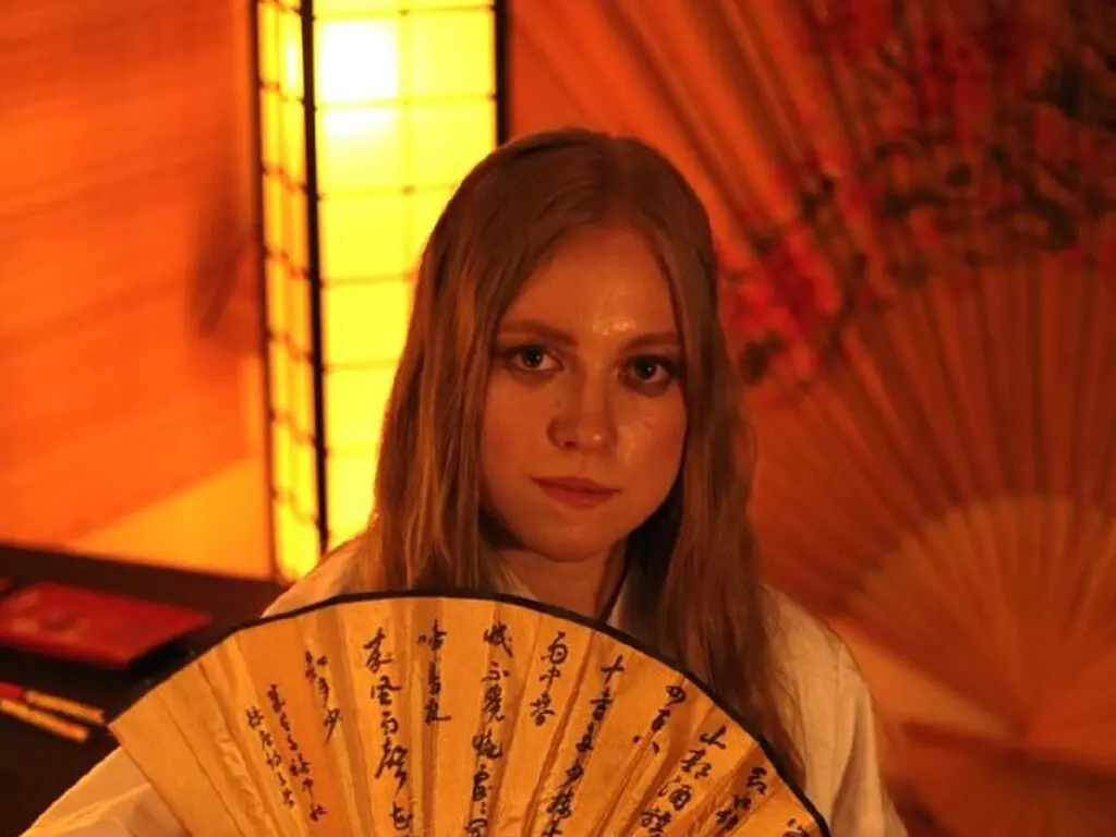 Blonde woman in white shirt holding a traditional Japanese fan in a warm, moody room with a glowing lantern.