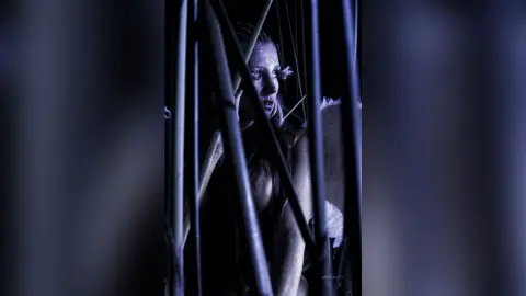 AndreaKalo poses behind dark vertical bars, showing her face and bare chest in a dim, moody setting.