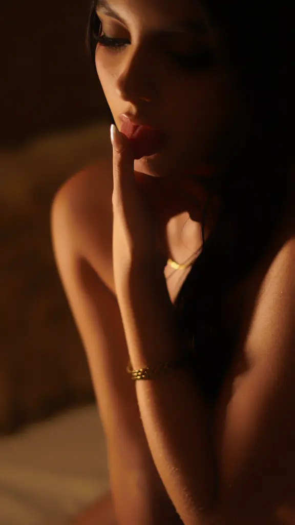 SamiLewis — Moody close-up of a woman with long dark hair wearing gold jewelry in warm, dim lighting.