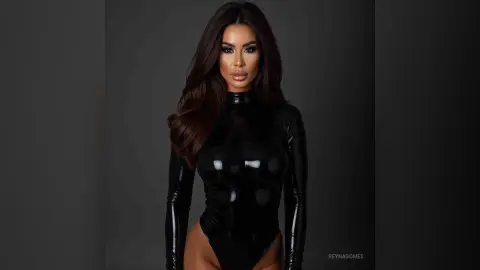 ReynaGomes poses in a shiny black long-sleeve bodysuit against a dark background, showing her long brown hair and curves.