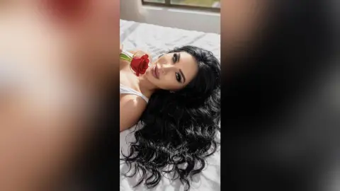 Beautiful woman with long dark hair holding a red rose while lying on white bedding in a bright room.