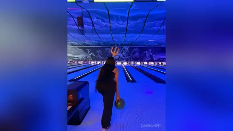 Woman in black outfit holding a bowling ball in a neon blue lit bowling alley with mural art.
