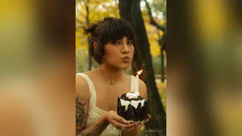 AlexiaVitali with dark hair and tattoos holds a small birthday cake with a lit candle in an outdoor setting.