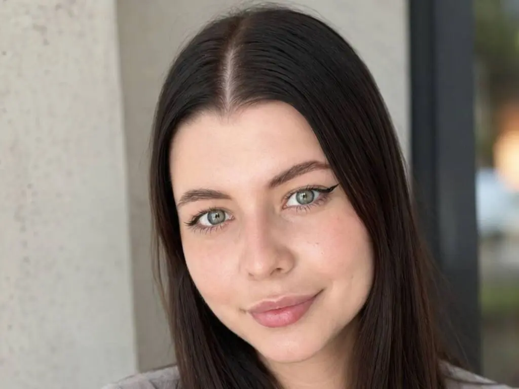 Close-up portrait of a smiling young woman with long dark hair and winged eyeliner in a natural outdoor setting.