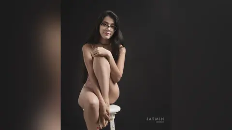 JessieLiu sits on a white stool wearing a light pink bodysuit, hugging her knees with long dark hair falling over her shoulders.
