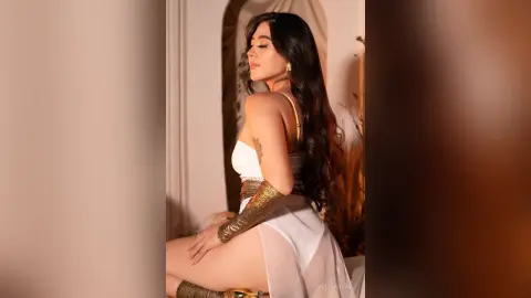 SofiCarvalo poses sideways in a white sheer outfit, showing her long dark hair and gold gloves against a light background.