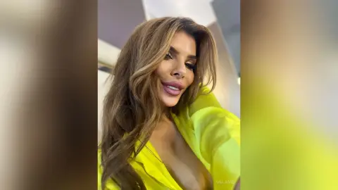 Glamorous woman with long wavy brown hair wearing a bright neon yellow outfit in a soft indoor setting.