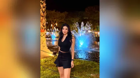 Elegant woman in a black sleeveless outfit posing by a glowing blue fountain in a nighttime garden setting.