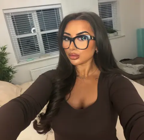 Young woman with long dark hair and black glasses wearing a brown long-sleeve top in a bright bedroom.