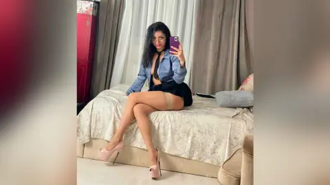 VeronicaQuinn — Woman in blue button-down shirt and black skirt sitting on a bed taking a mirror selfie in a cozy bedroom.