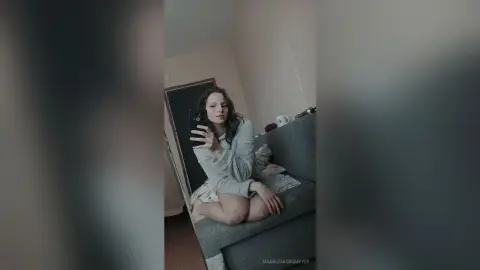 Young woman in a cozy white robe taking a selfie while sitting on a grey sofa in a dimly lit room.