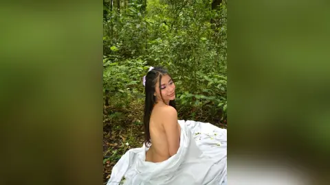 AmeliaStell poses naked in a lush green forest, wrapped in a white sheet that drapes over her lower body and back.