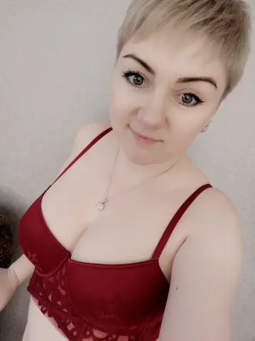 Smiling woman with short blonde hair wearing a red lace lingerie bra in a bright indoor setting.