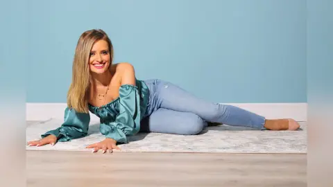 Gia smiles while lying on a light rug in light blue jeans and a teal off-the-shoulder blouse.