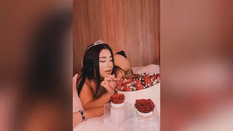 Young woman in a tiara lying on a bed with red roses in a romantic indoor setting.