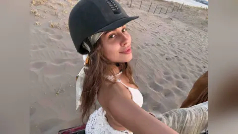 EvelynMills smiles while wearing a black helmet and white lace top, sitting outdoors near a sandy beach area.