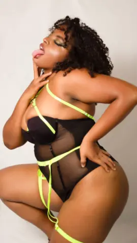 Curvy Black woman in black and neon yellow lingerie posing playfully against a white background.