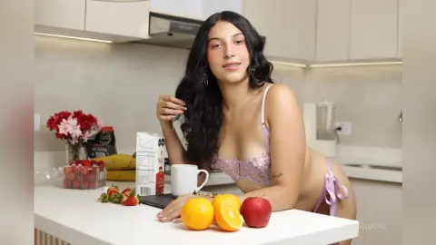 Smiling woman in lavender lace lingerie sitting at a kitchen counter with fresh fruit and flowers.