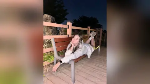 Young woman in a white long-sleeve shirt relaxing on a wooden park bench at night.