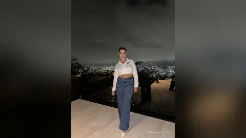 Stylish woman in a white long-sleeve crop top and blue jeans standing on a balcony overlooking a city at night.