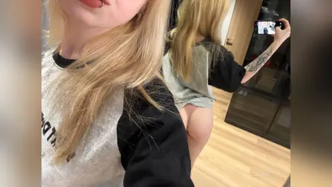 Blonde woman in a grey graphic t-shirt taking a mirror selfie in a modern room.