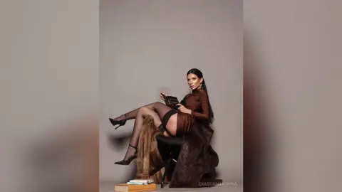 Elegant woman in a brown long-sleeve dress sitting on a chair against a neutral studio background.