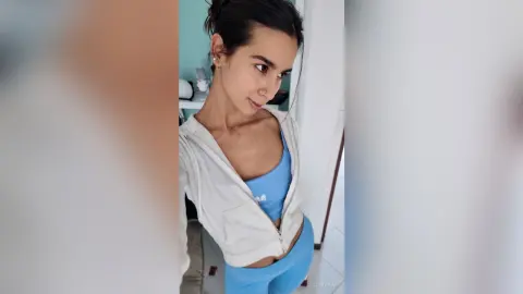 Young woman in blue yoga outfit and white hoodie posing in a bright room with a relaxed mood.