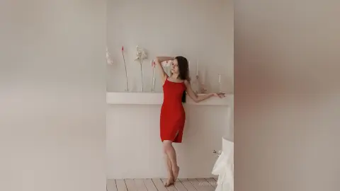 IngridBeckton poses in a red sleeveless dress with a side slit, standing against a white wall and leaning on a white shelf.