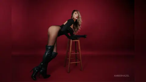 AmberHazel poses on a wooden stool wearing a black bodysuit and long black gloves against a red background.