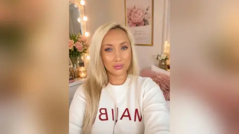 Blonde woman in a white sweatshirt smiling in a cozy, brightly lit bedroom setting.