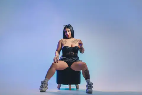 Tattooed woman in black lace lingerie sitting on a stool against a blue studio background.