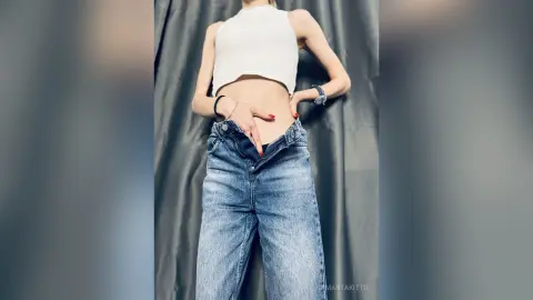 Woman in white crop top and blue denim jeans posing against a grey curtain backdrop.
