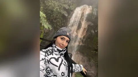 Smiling woman in a white patterned winter jacket and grey beanie standing before a misty forest waterfall.