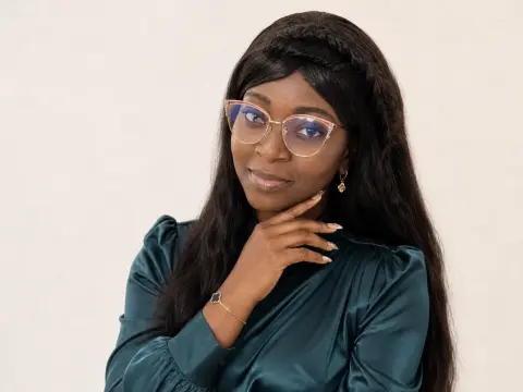 Smiling Black woman with glasses and dark hair wearing a dark green satin blouse in a bright studio setting.
