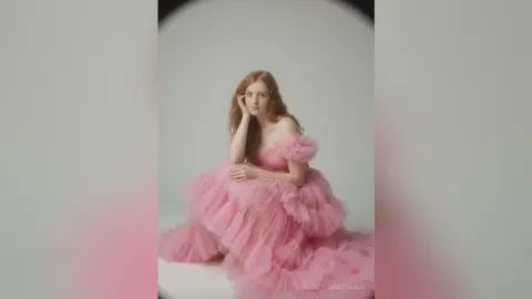 Redheaded woman in a voluminous pink tulle gown posing elegantly in a minimalist studio setting.