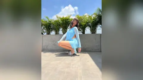 Young woman in a light blue summer dress posing on a sunny rooftop garden with palm trees.
