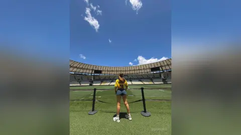 Young woman in a yellow t-shirt and denim shorts standing on a green field at a large outdoor stadium under a blue sky.