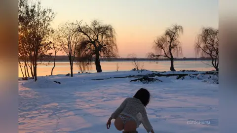 Woman in a white knit sweater sitting in a snowy landscape by a lake during a peaceful winter sunset.