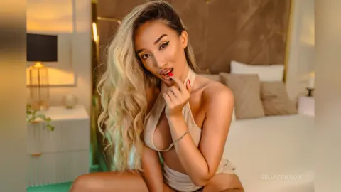 Glamorous blonde woman in a beige halter top posing in a cozy, warm-lit bedroom setting.