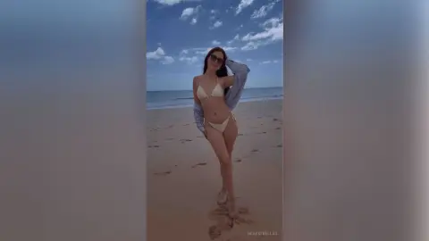 Stylish woman in a white bikini and sunglasses posing on a sunny beach under a blue cloudy sky.