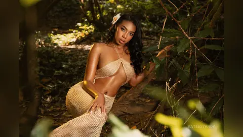 Beautiful Black woman in a gold crochet bikini outfit posing in a lush, sunlit tropical forest setting.