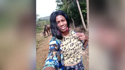 Smiling woman in a patterned cream top and blue floral cardigan standing outdoors near a horse in a rural field.
