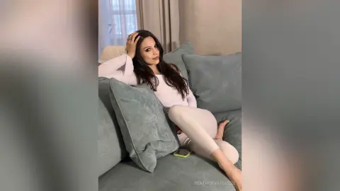 Relaxed woman in a beige lounge outfit reclining on a grey sofa in a bright, cozy living room.