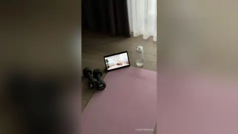 ChristineDavis poses on a pink yoga mat next to black dumbbells and a water bottle in a bright room with dark curtains.