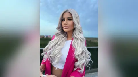 Stylish woman with long wavy silver hair wearing a pink blazer and white top outdoors under a blue sky.