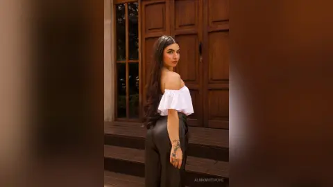 AlmaWhitmore poses outdoors in black pants and an off-the-shoulder white top, looking back over her shoulder with long dark hair.