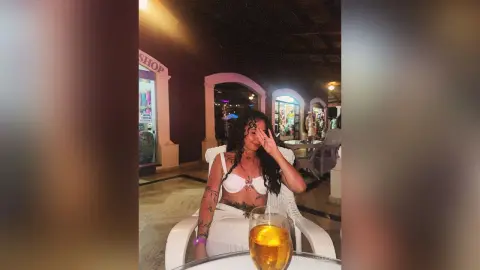 RossiNoleen sits outdoors at night wearing a white bra and white skirt, holding her hand to her face near a glass of amber liquid.