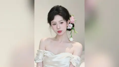Elegant woman in an off-the-shoulder cream satin dress with pink floral hair accessories in a soft aesthetic setting.